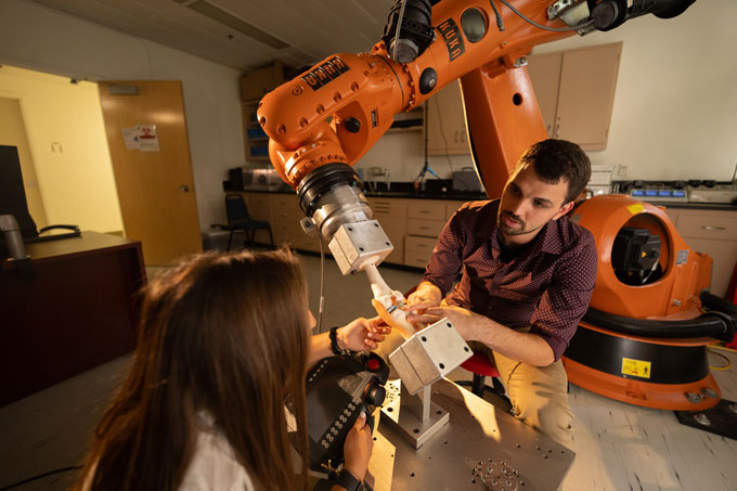 Two people sit on either side of a human knee model that is grasped by a large orange robotic arm